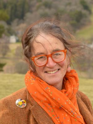 Photograph of Lesley Morrison. Lesley is wearing an orange scarf and a brown jacket. She is standing outside in front of some tr