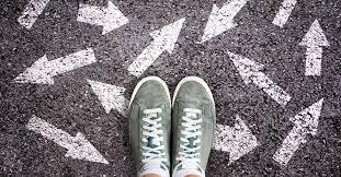 Image of feet standing on a pavement with white arrows on the ground pointing in many different directions