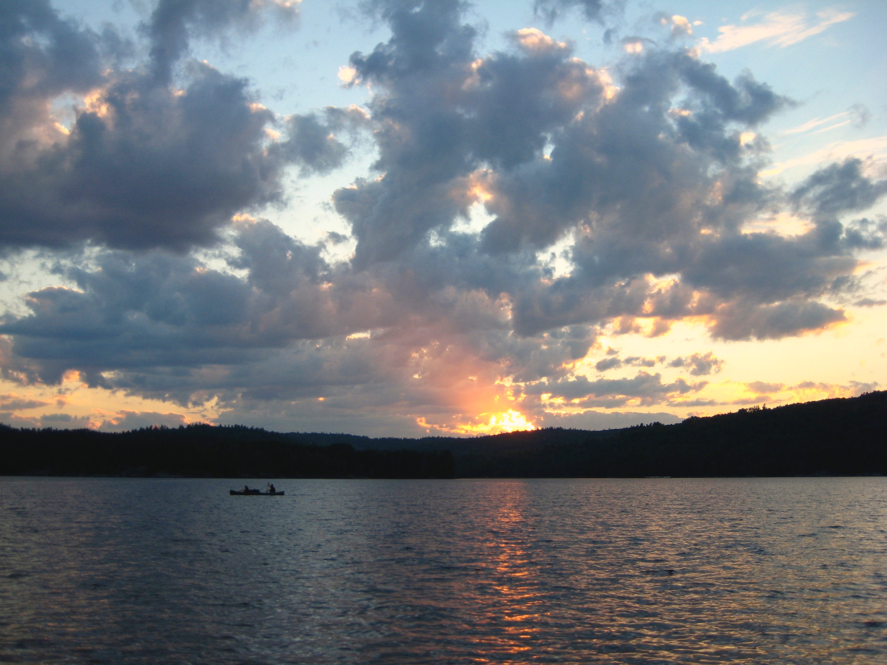 Photograph of Grand Lake, Ontario. There is a small boat on the water, beyond the water there is the silhouette of the shoreline and above their is a sunset behind grey clouds. 