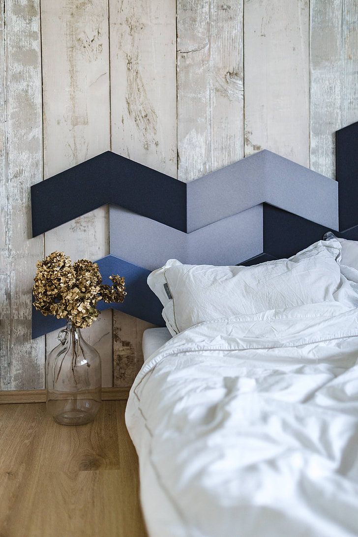 Photograph of a cosy bed, with white sheets. On the wooden floor beside the bed there is a vase with dried flowers in it. At the top of the bed is a headboard with different blue wooden pieces. 