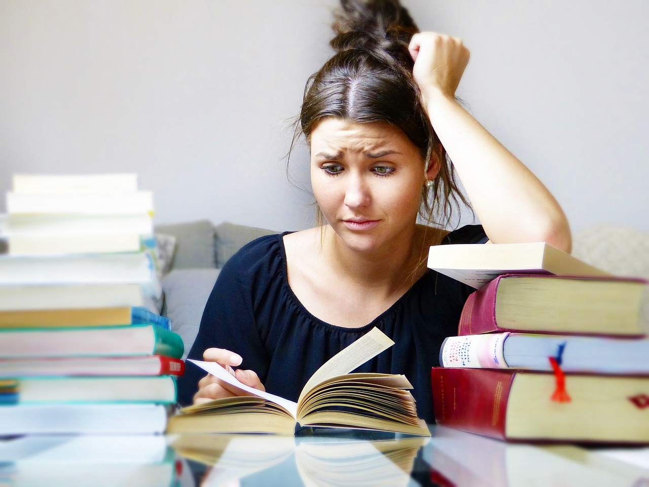 Photograph of a woman wearing a black shirt, sitting at a table. She is reading a book with lots of books pilled around her. 