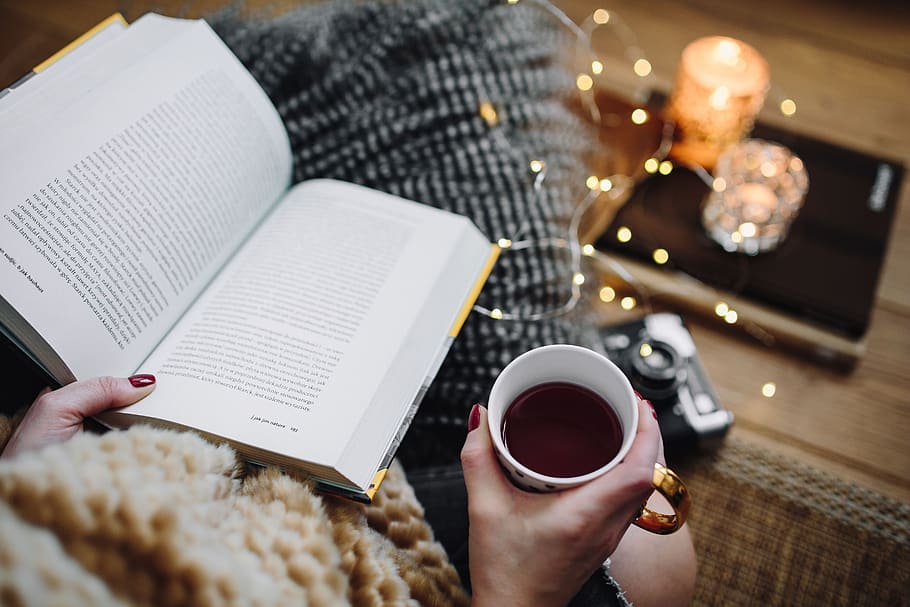 Photograph of a person drinking coffee while reading in a book in a cosy setting 