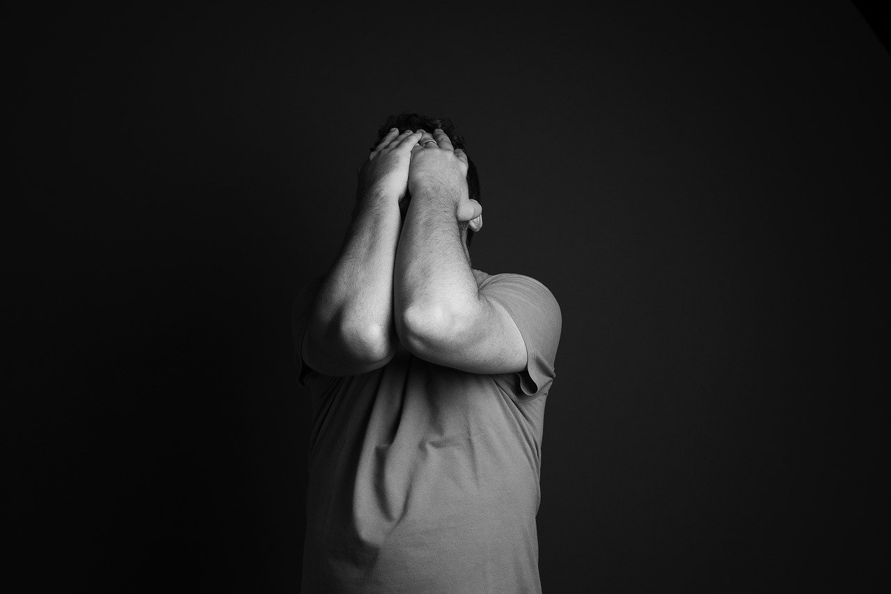 Black and white photograph showing a man with his head in his hands. He is standing in front of a black wall 