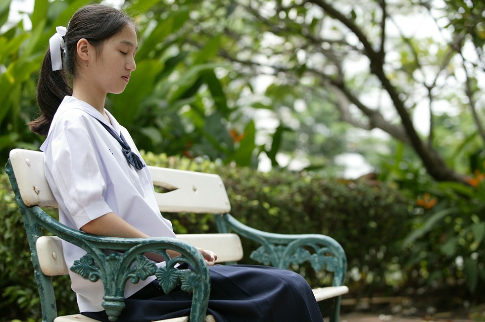 Photograph of a young woman, sitting on a bench in a garden with her eyes closed. 