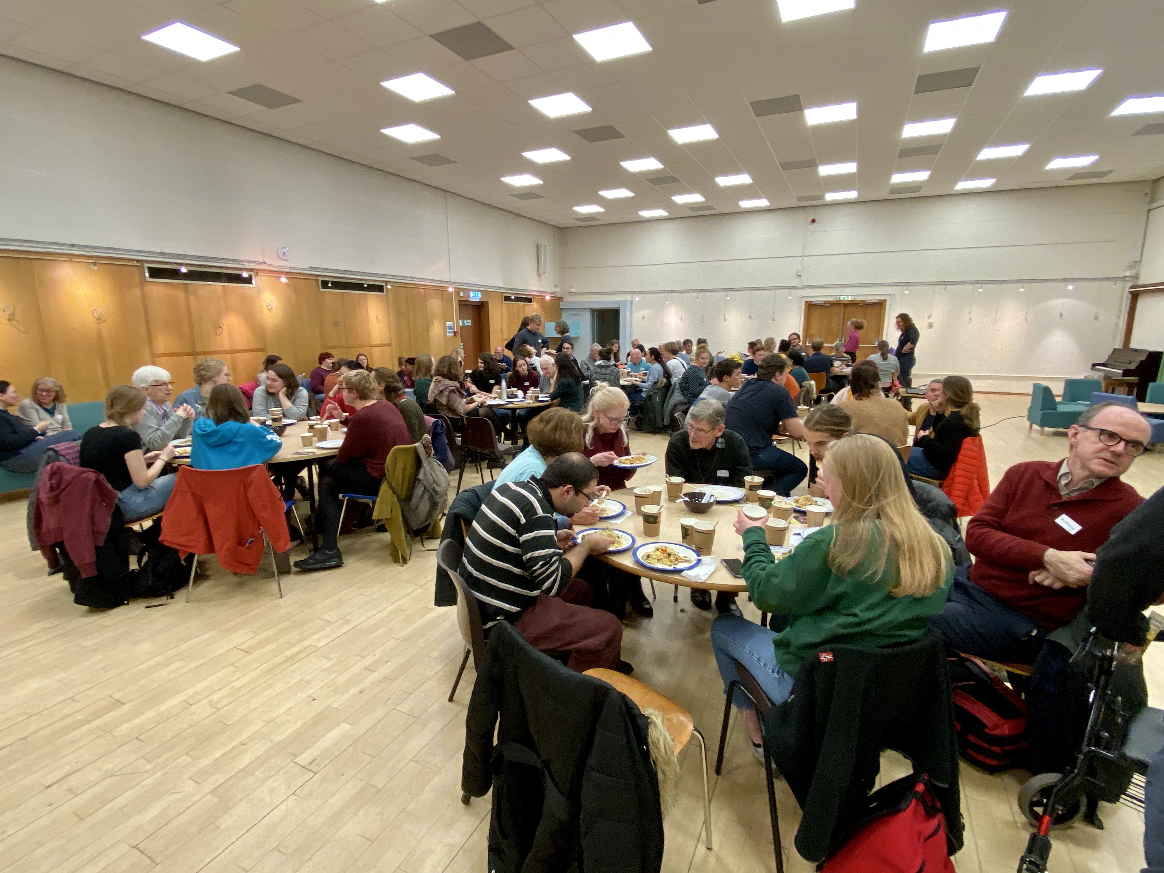 Photograph of a meal being held in the auditorium in The Chaplaincy Centre. There are groups of people sitting talking at tables