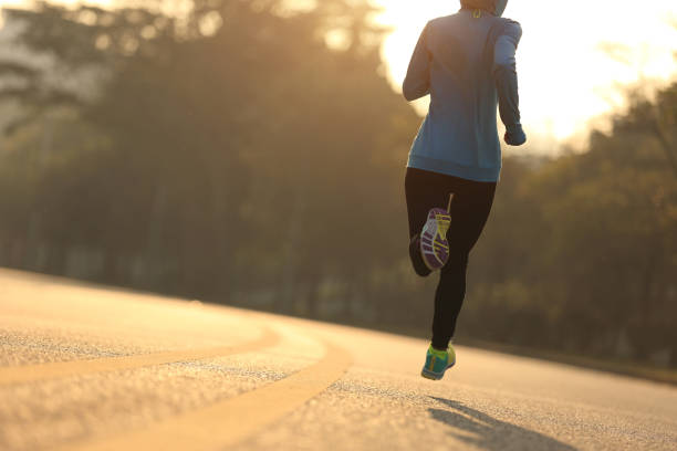 Photograph of a woman running on a road away from the camera. There are trees in the background and the sun setting