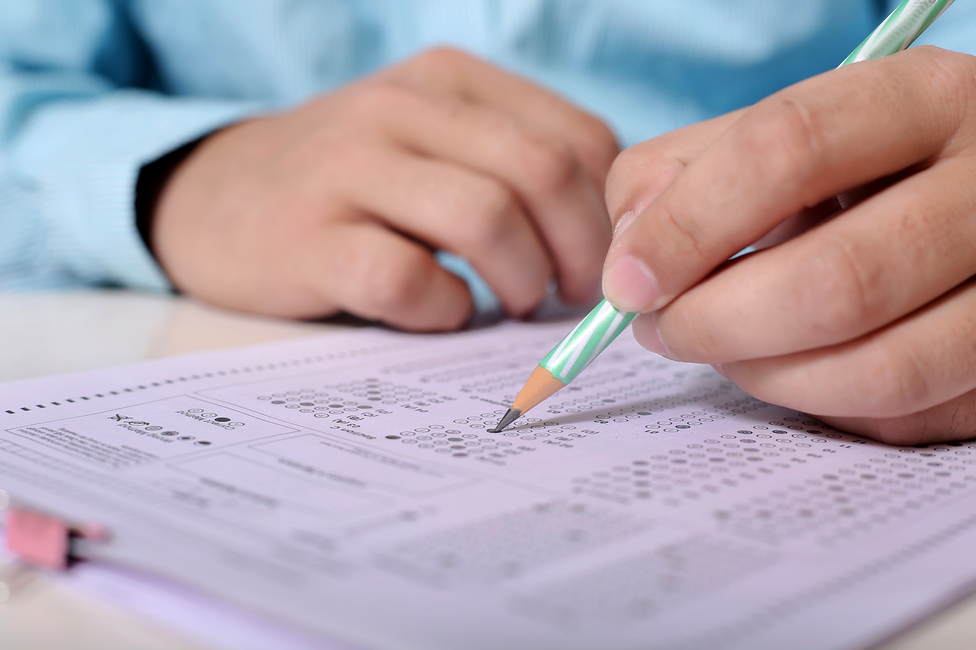 Photograph of a person's hands on a table answering exam questions. The person is holding a pencil and marking his answers on the paper in front of him.