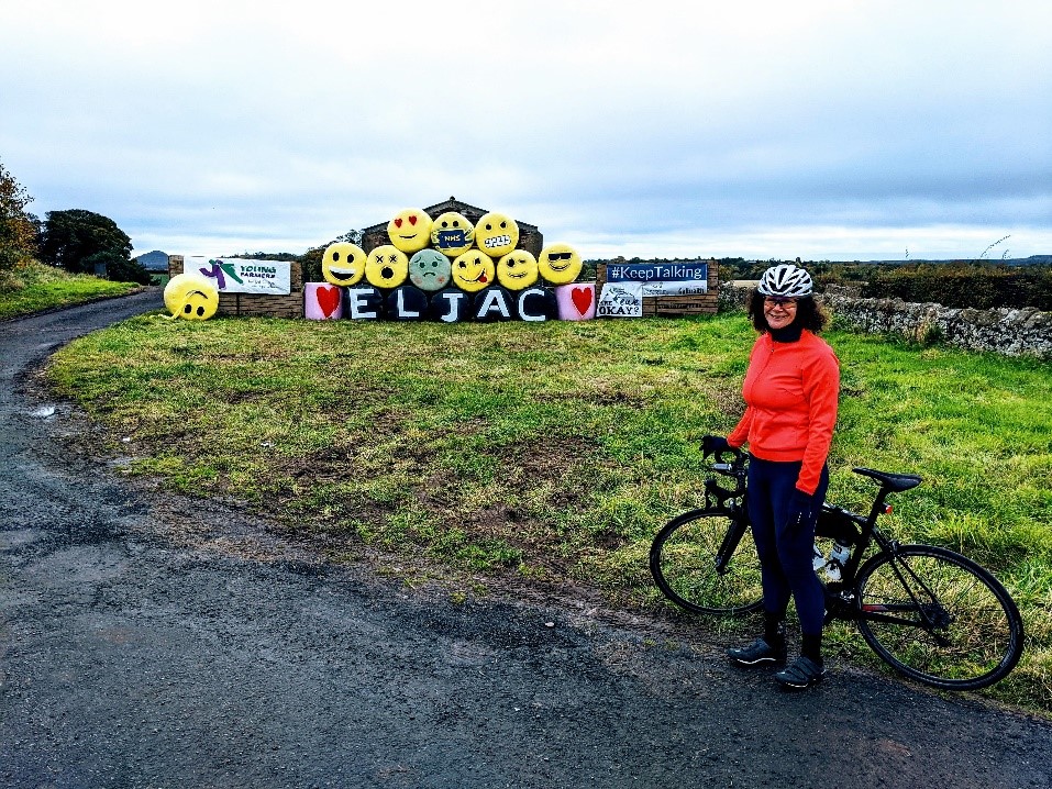 Photograph of Harriet Harris, University Chaplain, out for a ride on her bike. In the background there is covered hay stacked up. The hay covers are in the style of different emojis. 