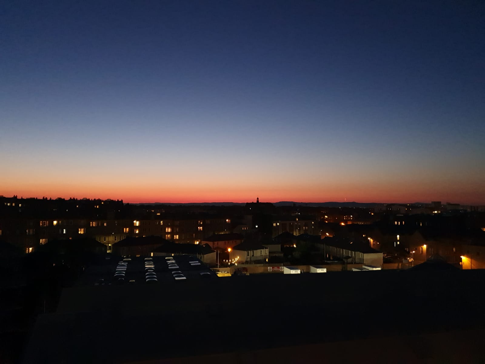 Photograph of a sunset over a city skyline. There are lights on in some of the houses and overhead the sky is darkening as the sun sets. 