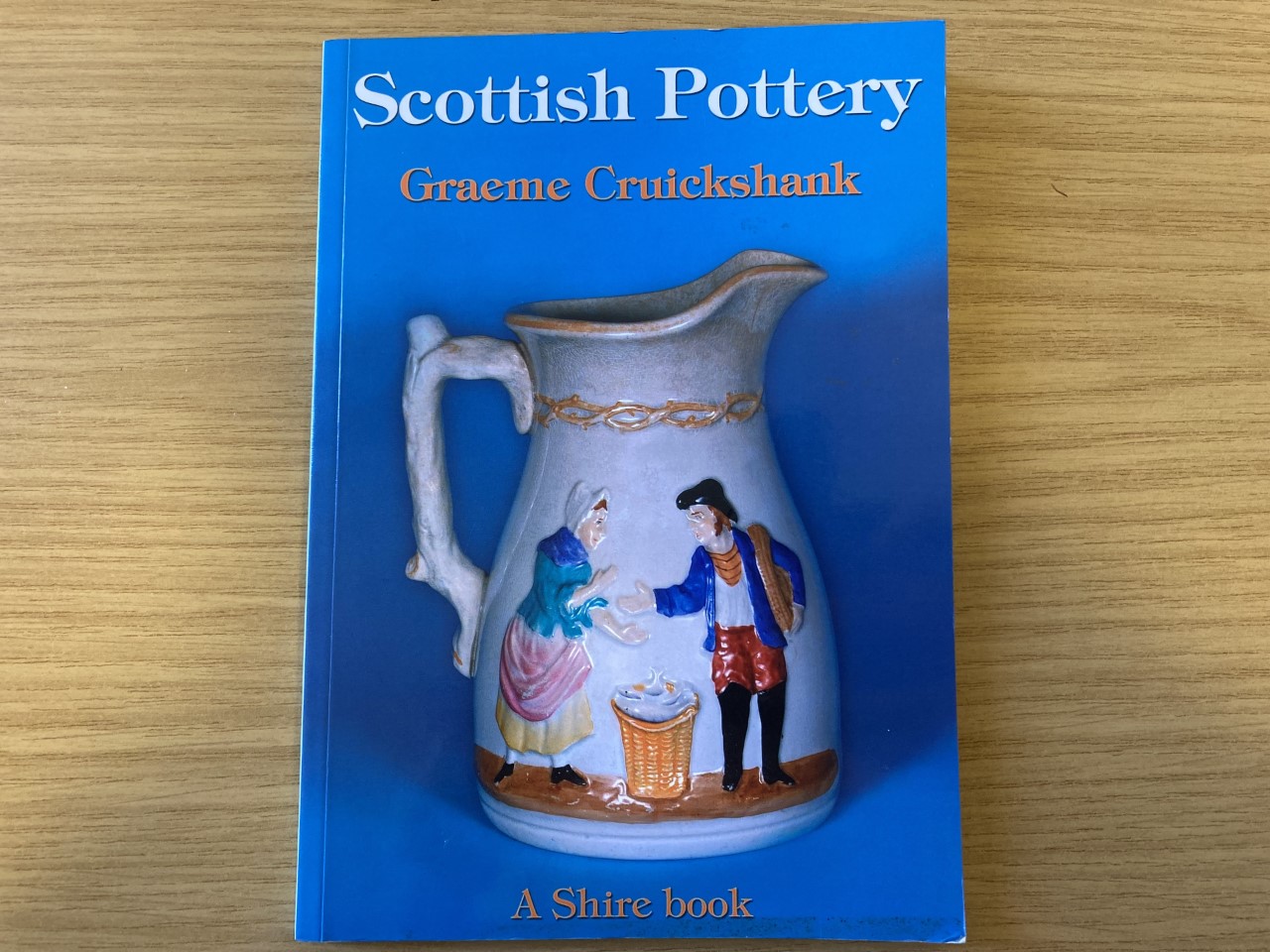 A photo of a book titled \"Scottish Pottery\" the front cover of the book shows a pot with two people enamelled on it.