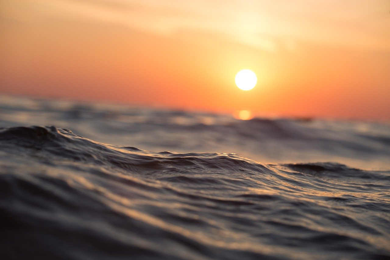 Photograph of the sea with small waves across the picture. In the background the setting sun, creating an orange sky.