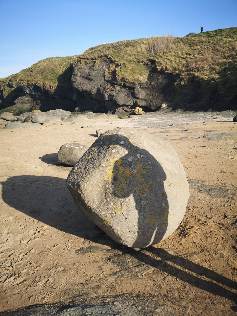 Shadow of a person on a rock, while another person walks along the cliff top in the distance