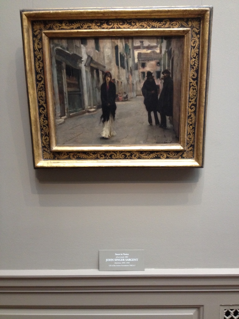 Photograph of John Sargent’s painting A Street in Venice hanging in a gallery. In the painting there is a woman walking through the streets of Venice.