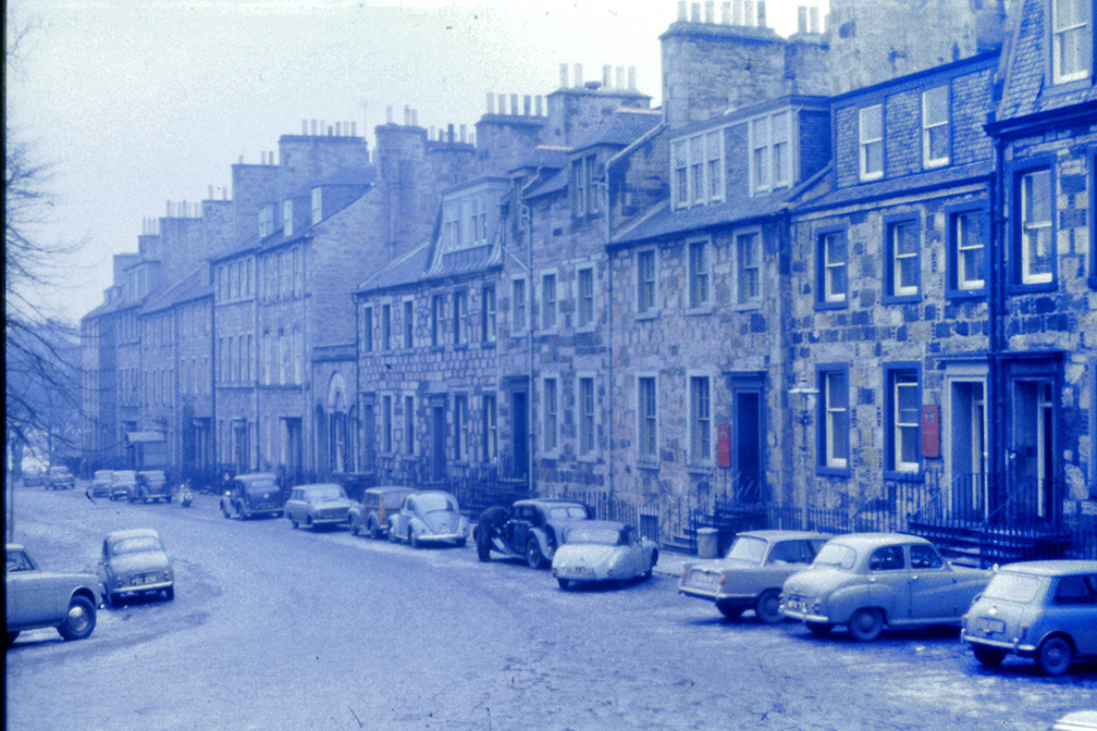 A black and white photograph of some of the buildings in George Square. There are old cars parked along the street. 