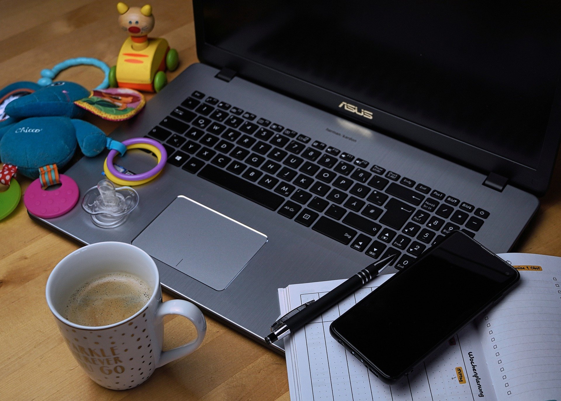 Photograph of a laptop on a desk, with a cup of tea at the side, notebooks with a phone on top of it. On the other side baby toy