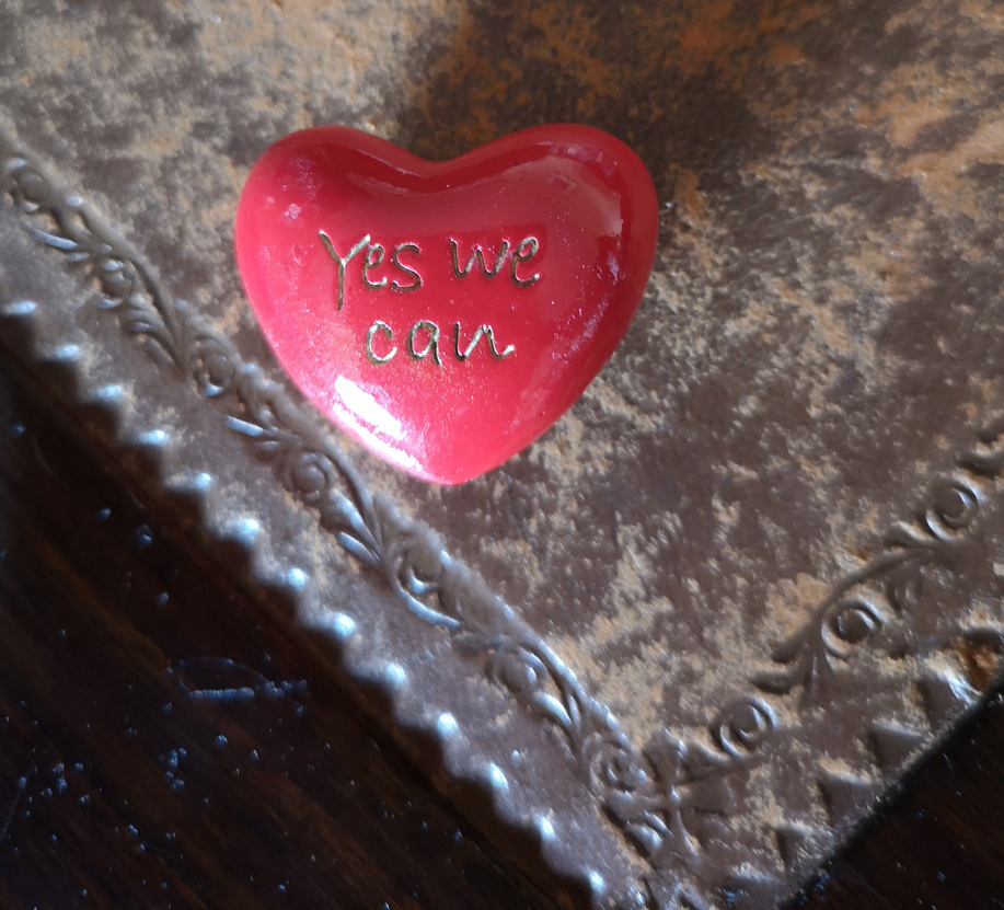 Photograph of a red stone in the shape of a heart with the words \"Yes We Can\" carved onto it. The red stone is sitting on the edge of a brown table. 